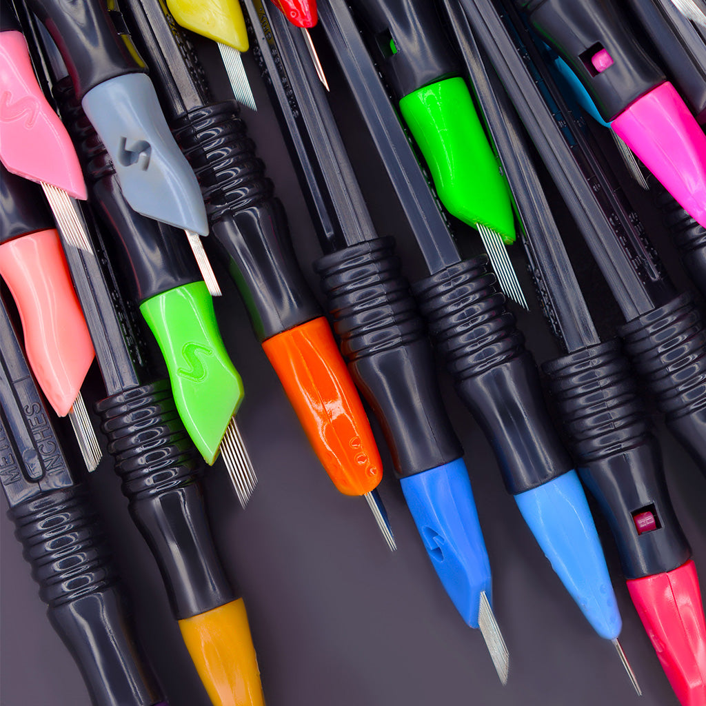 Close-up of several colorful SofTap permanent makeup click tip needles with black click stick handles arranged from top to bottom on a dark background.