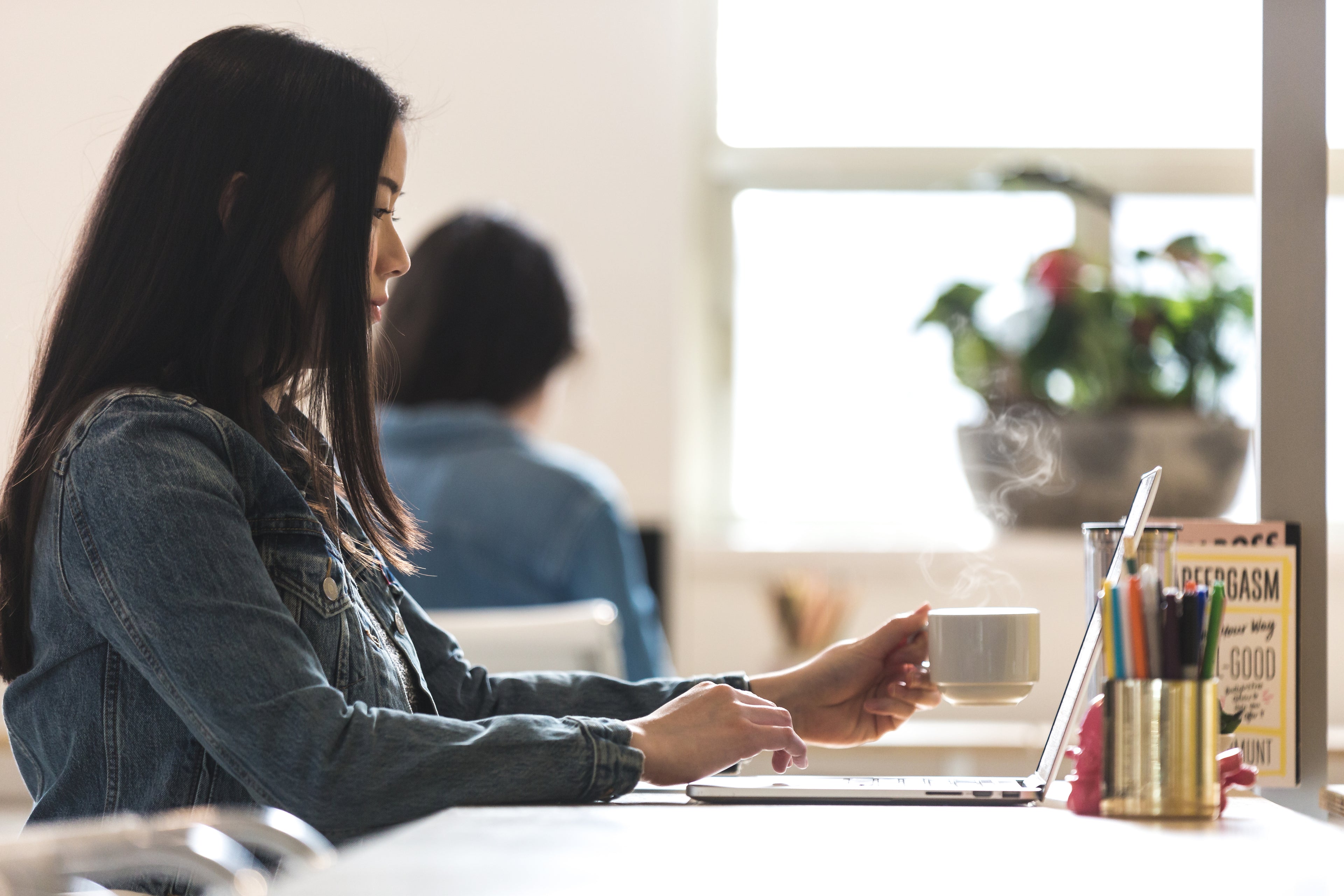 woman with dark hair in denim jacket working on a laptop with a white cup of coffee in a white office