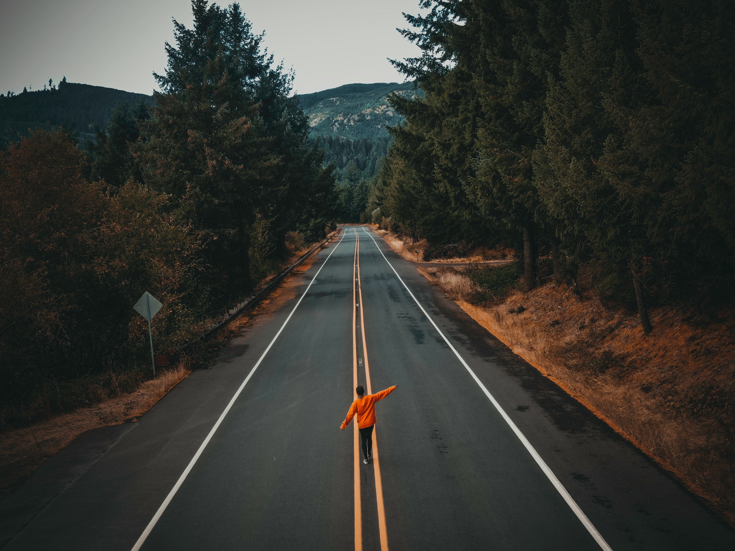 woman in orange sweater walking in the middle of a long empty two way road with a forest of trees on both sides with a green mountain in the middle