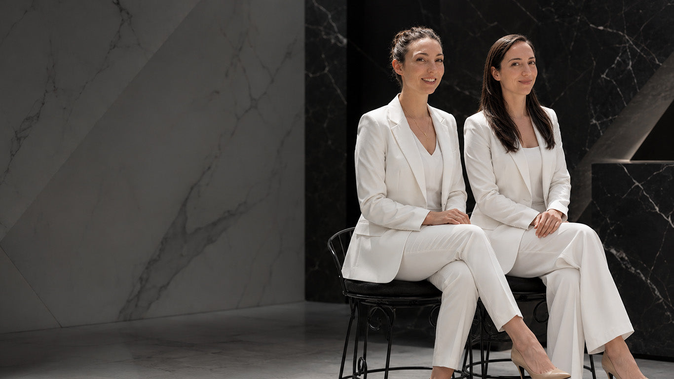 Two professional permanent makeup artists in white suits sitting on black stools against a dark marble wall.