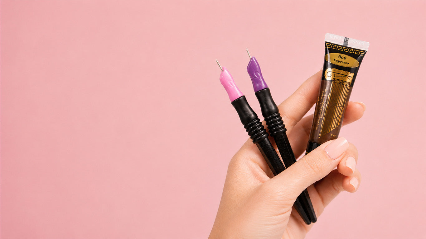 Hand holding two softap needles and one pmu pigment tube against a pink background