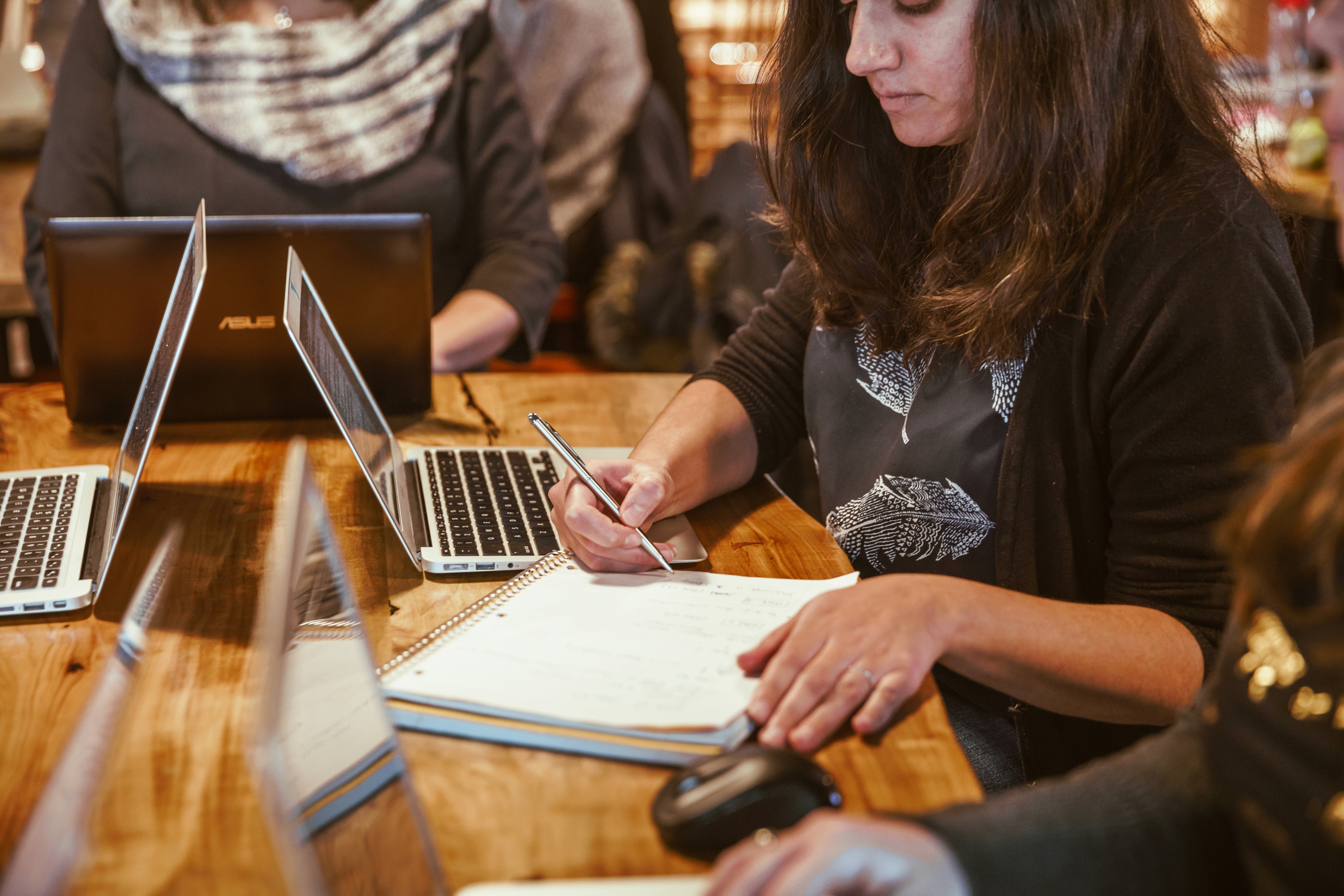 woman with dark hair and clothes working on laptop and taking notes in a cafe on a wooden table