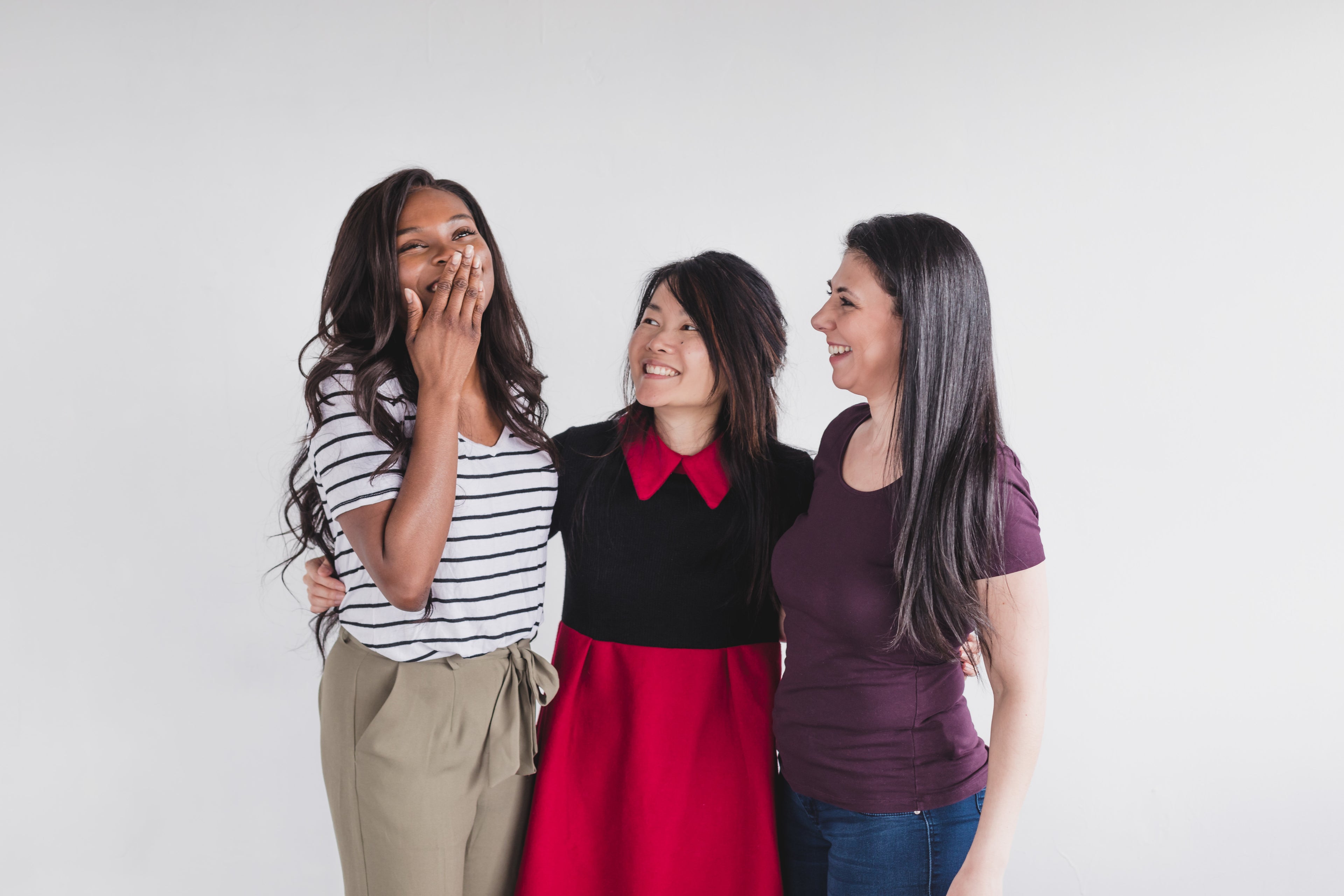 3 female friends with brown hair in a line and their arms around each other smiling with white background
