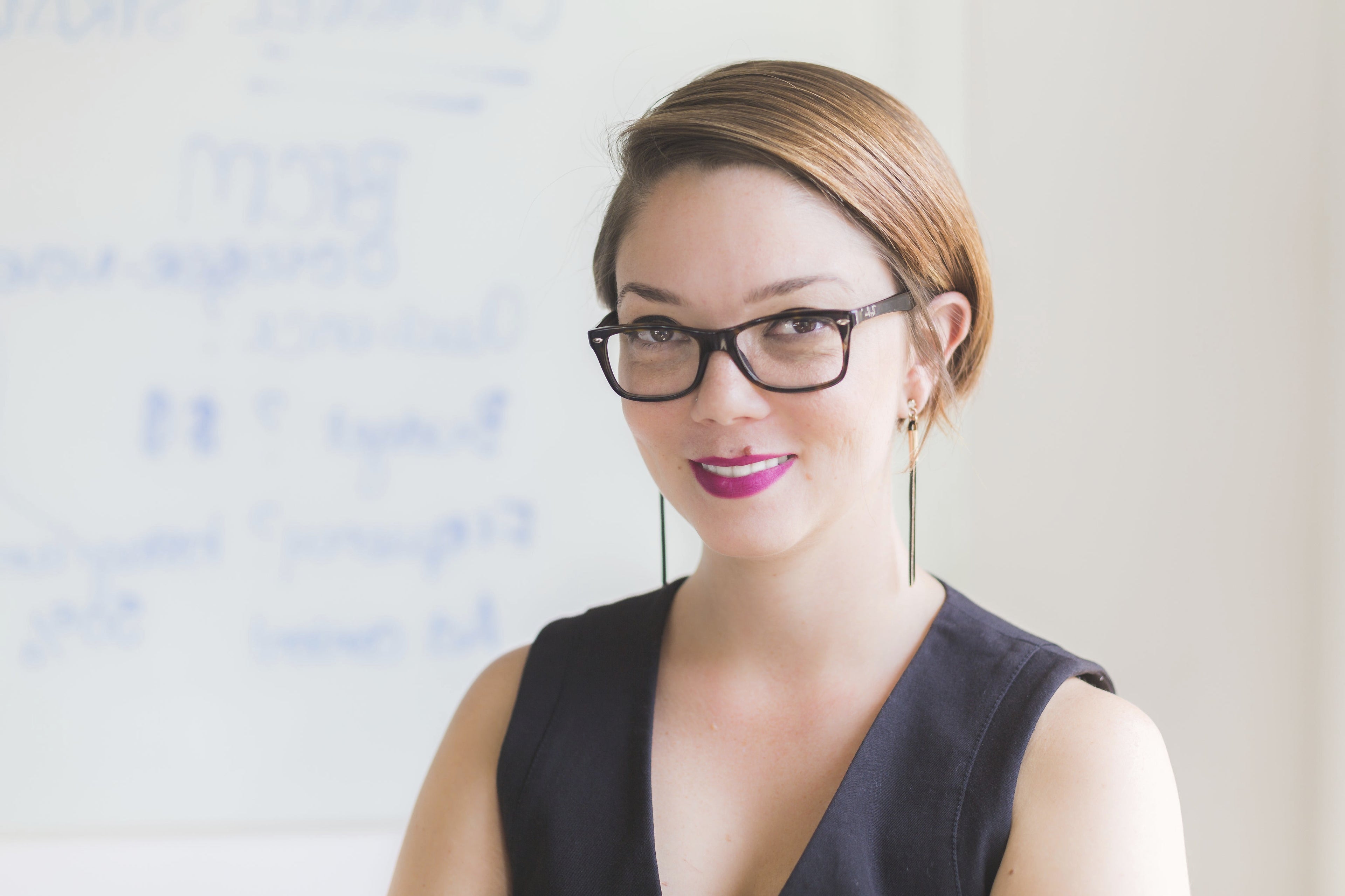 Woman wearing glasses and a black top in front of a whiteboard with text.